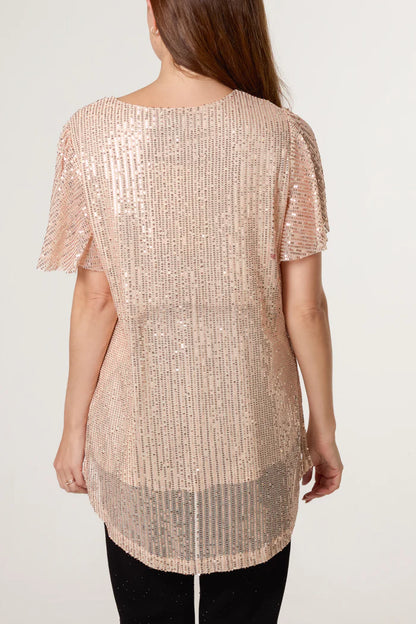 A person is seen from behind wearing a short-sleeved, blush pink sequin top—ideal for a festive wardrobe—teamed with black trousers, standing against a plain, light backdrop.