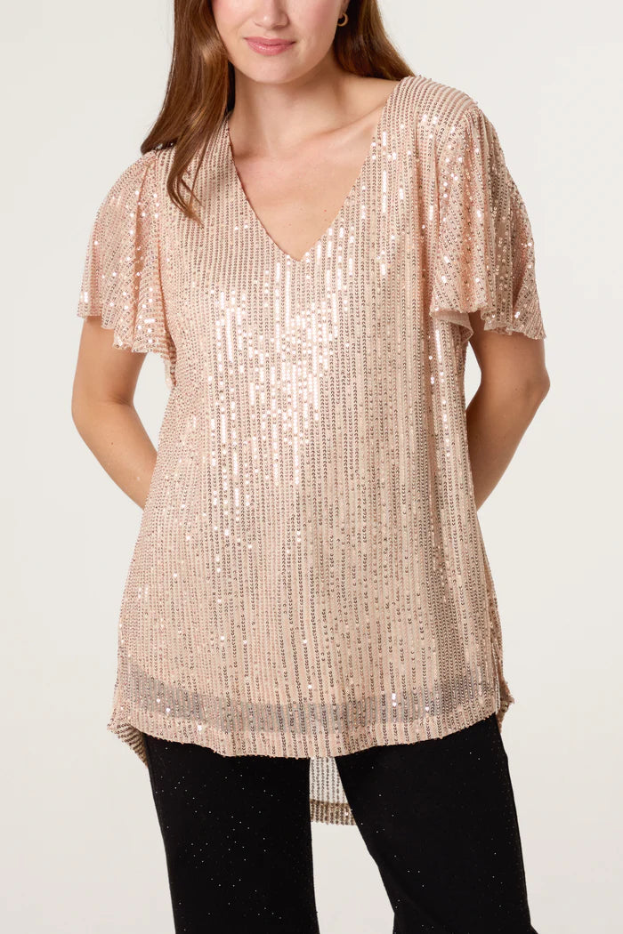 A person wearing a light pink, short-sleeved, sequin top with a V-neck, paired with black trousers, stands against a plain background—ideal for adding sparkle to your festive wardrobe.