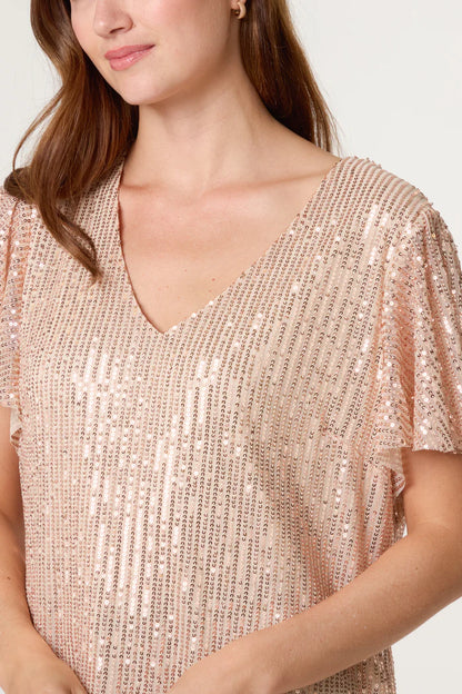 A woman wearing a light pink sequin top with short sleeves, vertical rows of shimmering sequins, and a V-neckline—ideal for adding a touch of glamour to your festive wardrobe.