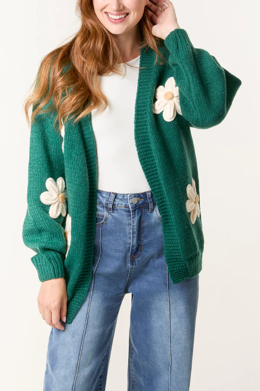 Woman wearing a sustainable green hand-knitted cardigan with large white flower appliqués, over a white blouse and high-waisted blue jeans.