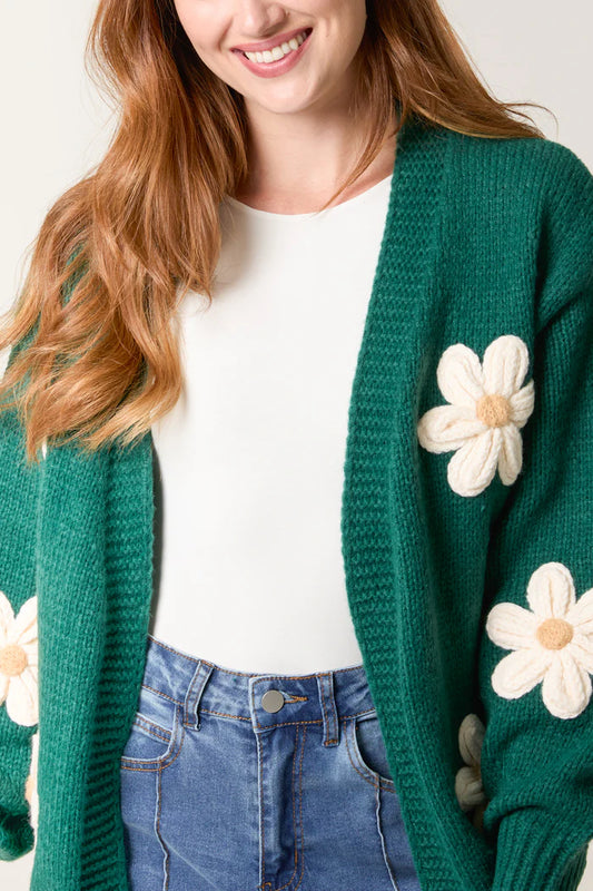 Woman wearing a green vegan knitted cardigan with white flower appliqués over a white top and blue high-waisted jeans, smiling.