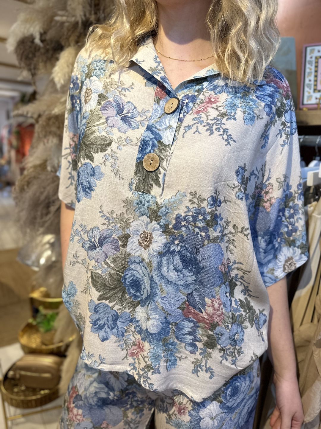 Woman dressed in a beige floral co-ord set with blue and green embroidered-style print; consists of a loose short-sleeved shirt with wooden buttons and wide-leg trousers.