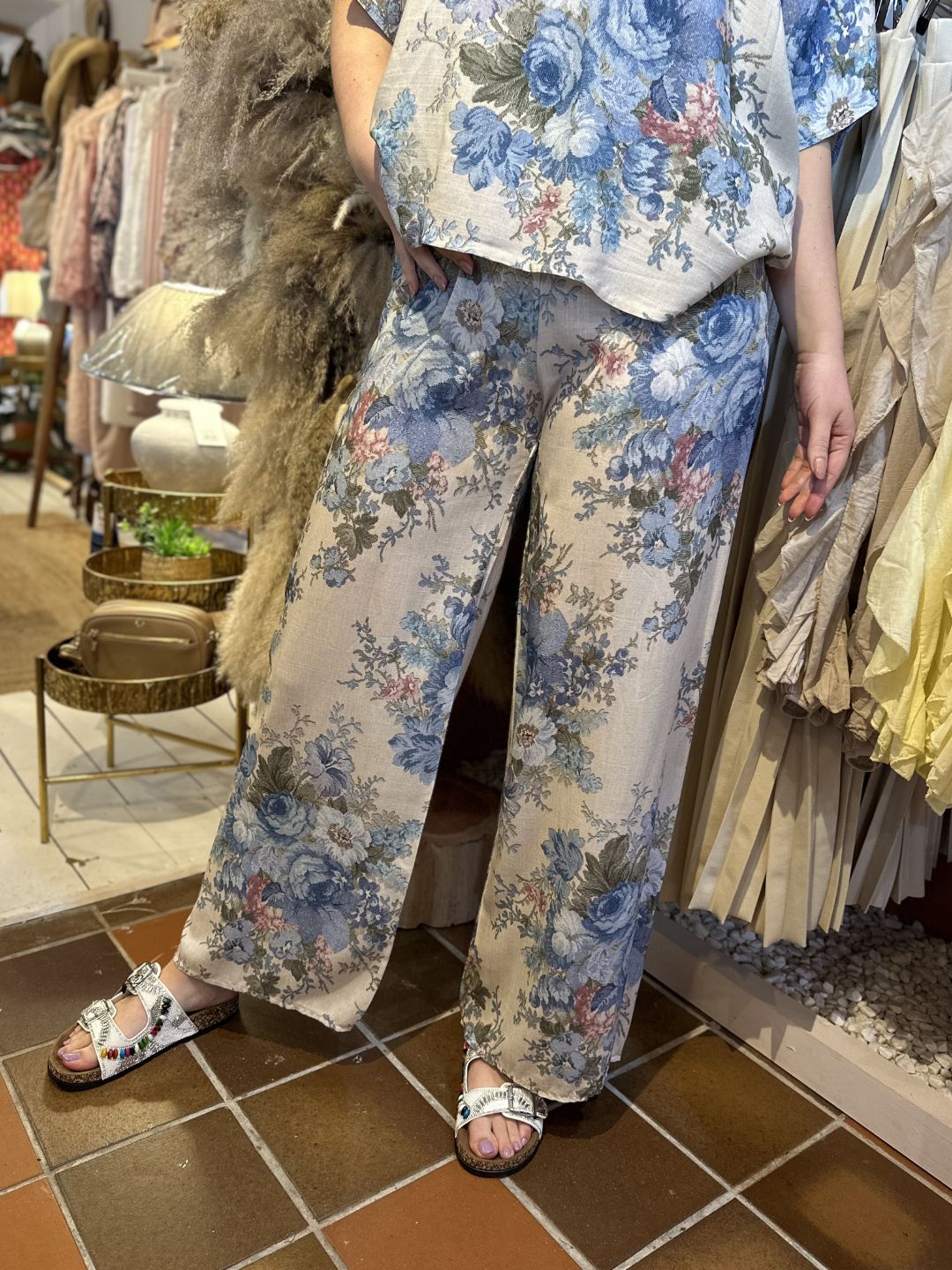 Person wearing a matching floral co-ord set with wide-leg trousers and white sandals, standing on a brown tiled floor in a shop filled with various textiles and decorative items in the background.