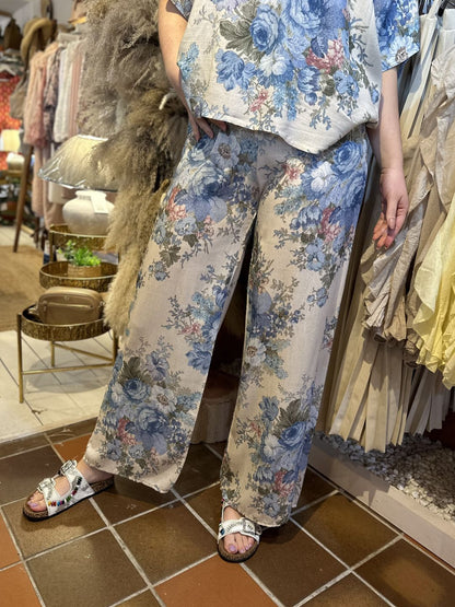 Person wearing a matching floral co-ord set with wide-leg trousers and white sandals, standing on a brown tiled floor in a shop filled with various textiles and decorative items in the background.
