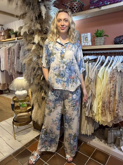 A woman stands in a boutique wearing a floral co-ord set with wide-leg trousers. She is surrounded by clothing rails, pampas grass, and stylish home décor items.