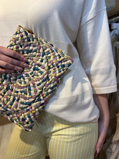 Person wearing a lightweight knitted white top and yellow striped trousers holds a multicoloured woven clutch bag against their side.