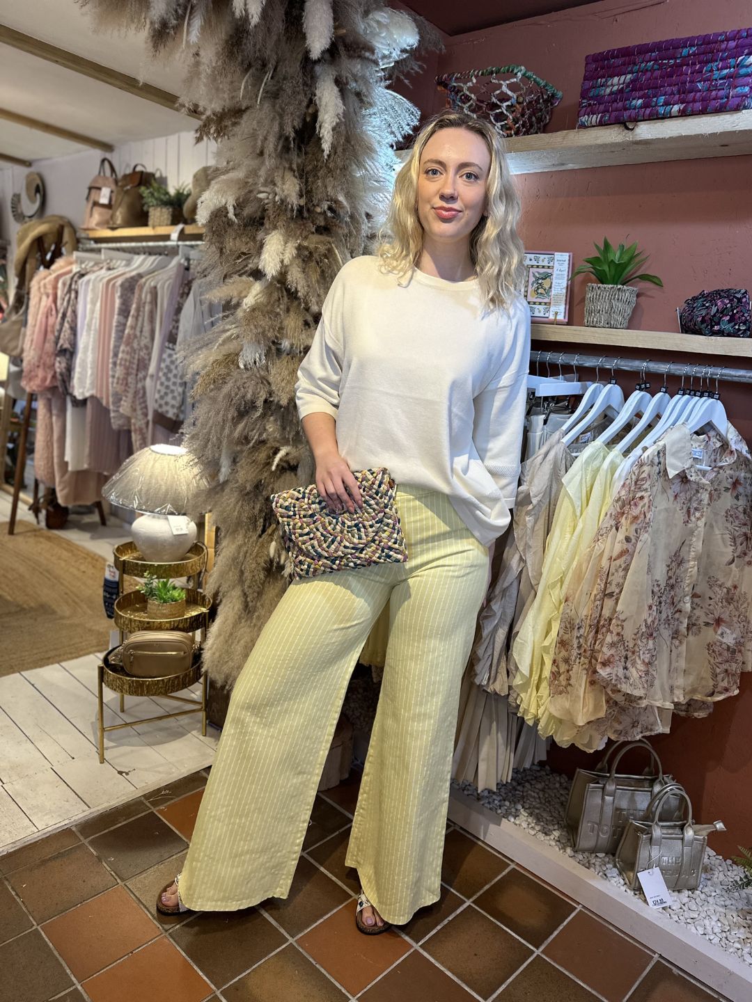 A woman stands indoors beside a clothing rail, dressed in a cream lightweight knit white top and yellow striped trousers, holding a patterned clutch. She is surrounded by hanging clothes and decorative plants.