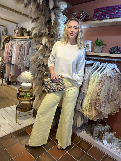 A woman stands indoors beside a clothing rail, dressed in a cream lightweight knit white top and yellow striped trousers, holding a patterned clutch. She is surrounded by hanging clothes and decorative plants.