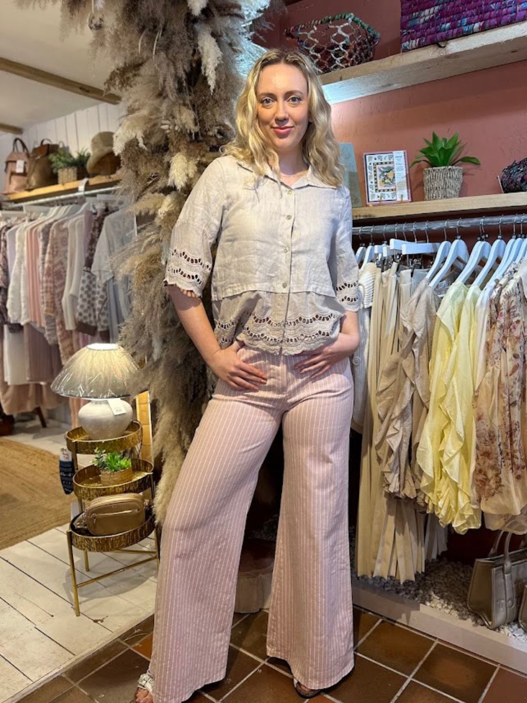 A woman stands in a boutique wearing a light beige broderie anglaise blouse with eyelet details and pink striped wide-leg trousers, surrounded by racks of clothing and decorative items.