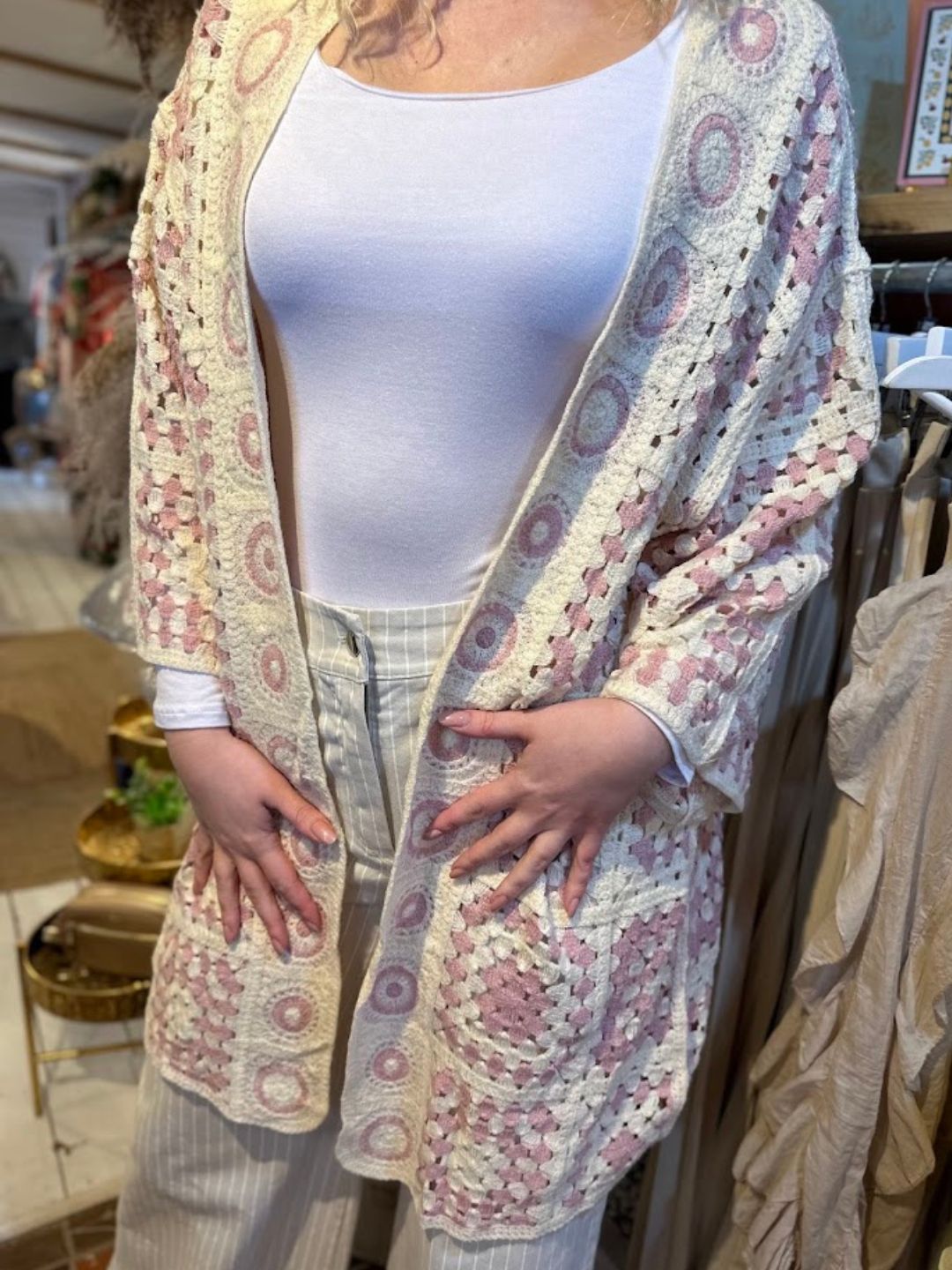 Person wearing a white top, light striped trousers, and a boho cardigan with pink and cream crochet patterns, standing indoors with hands resting on the cosy cardigan.