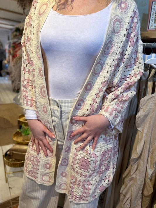 Person wearing a white top, light striped trousers, and a boho cardigan with pink and cream crochet patterns, standing indoors with hands resting on the cosy cardigan.