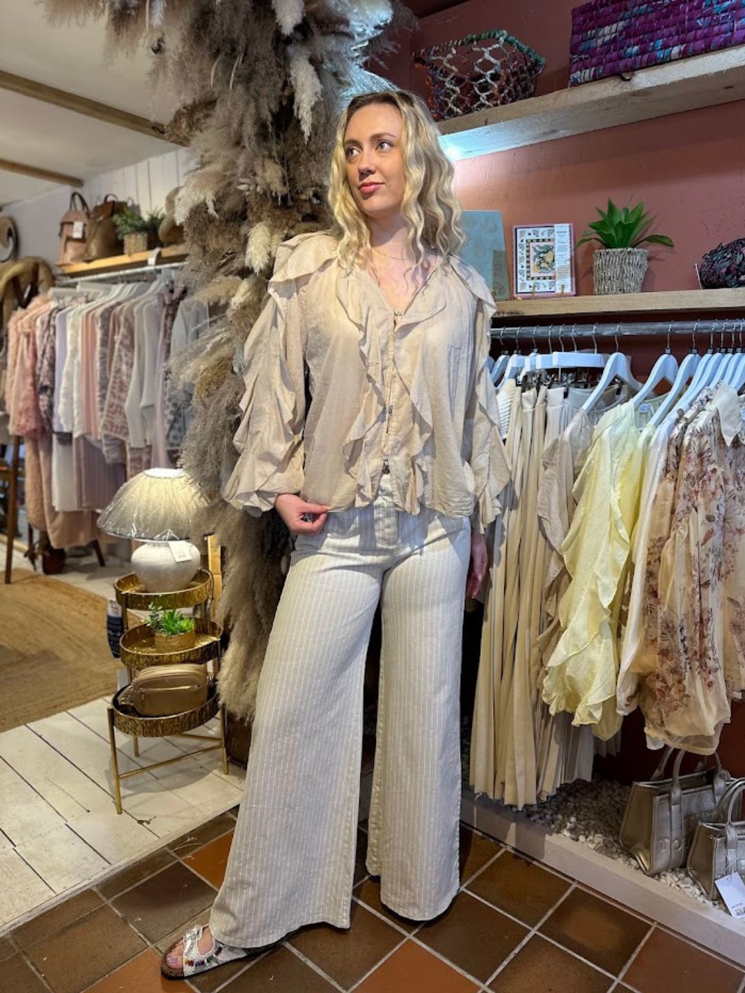 A woman stands in a boutique wearing a ruffle-front blouse, wide-leg striped trousers, and sandals, embodying feminine elegance among clothing rails and stylish décor in the background.