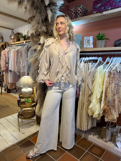 A woman stands in a boutique wearing a ruffle-front blouse, wide-leg striped trousers, and sandals, embodying feminine elegance among clothing rails and stylish décor in the background.