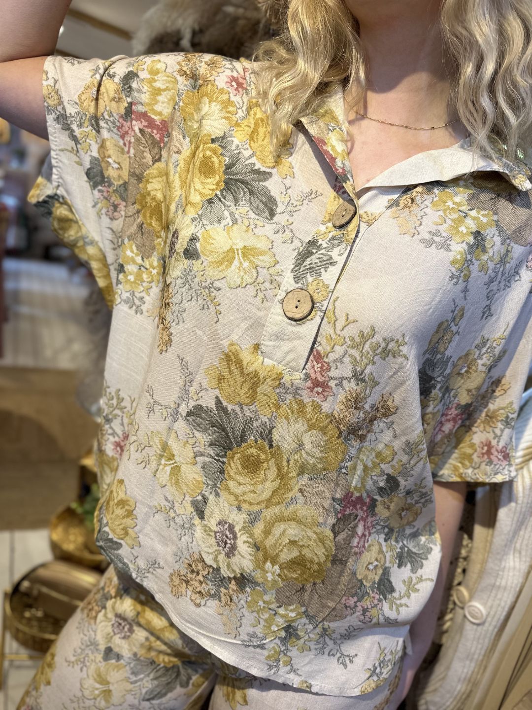 Person wearing a short-sleeved, button-up shirt and matching wide-leg trousers featuring a yellow and green floral pattern. Only the upper body and part of the face are visible.