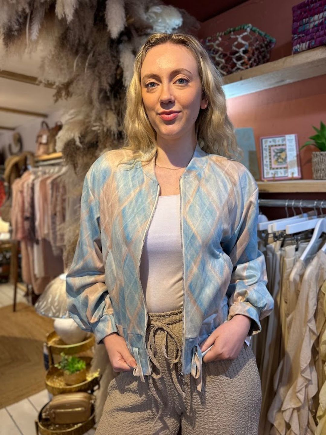 A woman in a light blue and beige checked bomber jacket over a white top stands indoors in a clothing shop, with racks of clothes and decorative items visible in the background.