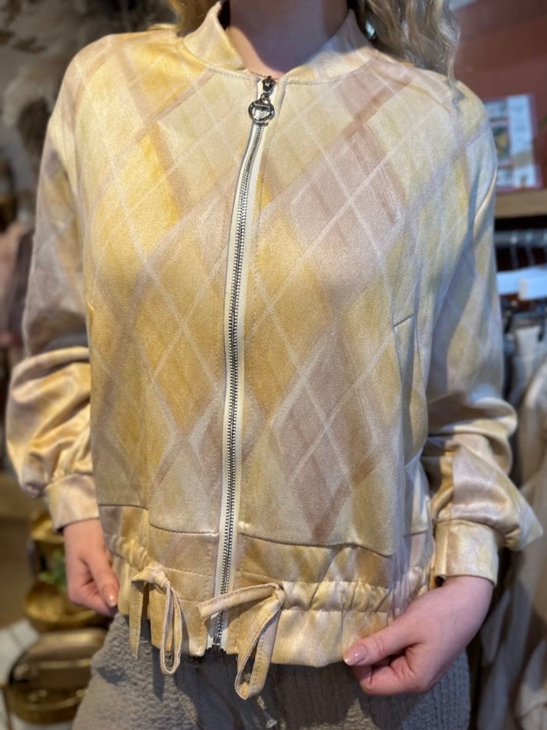 A person wearing a light yellow and beige checked bomber jacket with a zip-up front and drawstring hem, standing indoors with hands in their pockets.