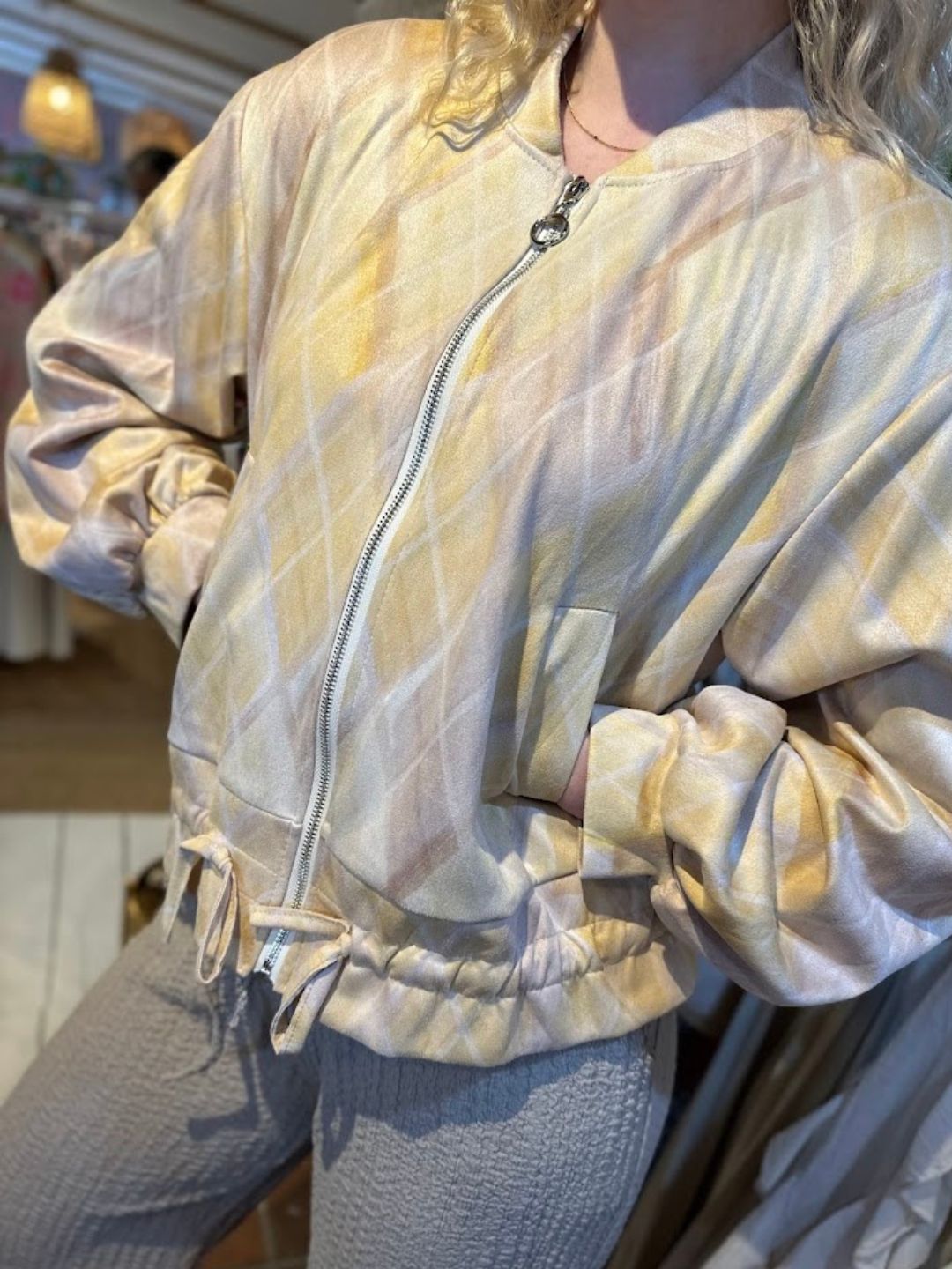 A person wearing a light yellow and beige colour-block jacket with ruched sleeves and textured light grey trousers, standing indoors.