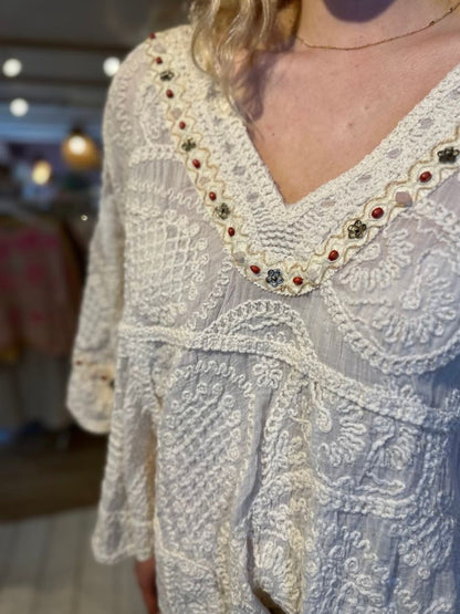 A close-up of a person wearing a white, vintage-inspired top with embroidered floral details along the neckline and sleeves. The background is gently blurred.
