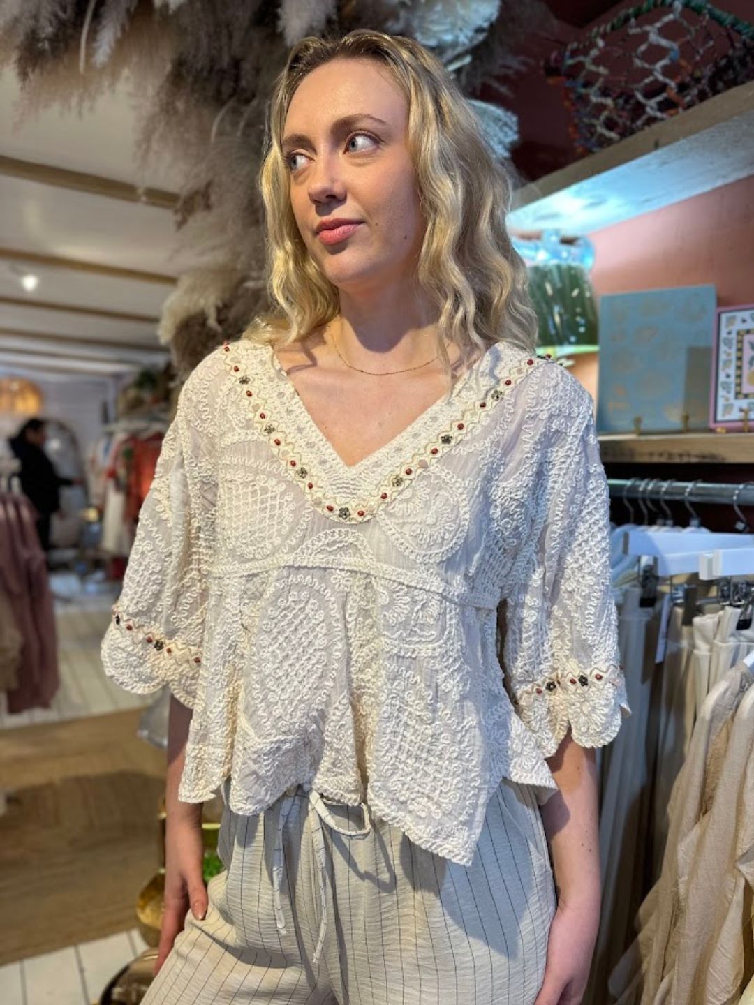 A woman stands indoors wearing a vintage-inspired, white bohemian blouse with scalloped sleeves and light-coloured, striped trousers. Clothing rails and décor are visible in the background.