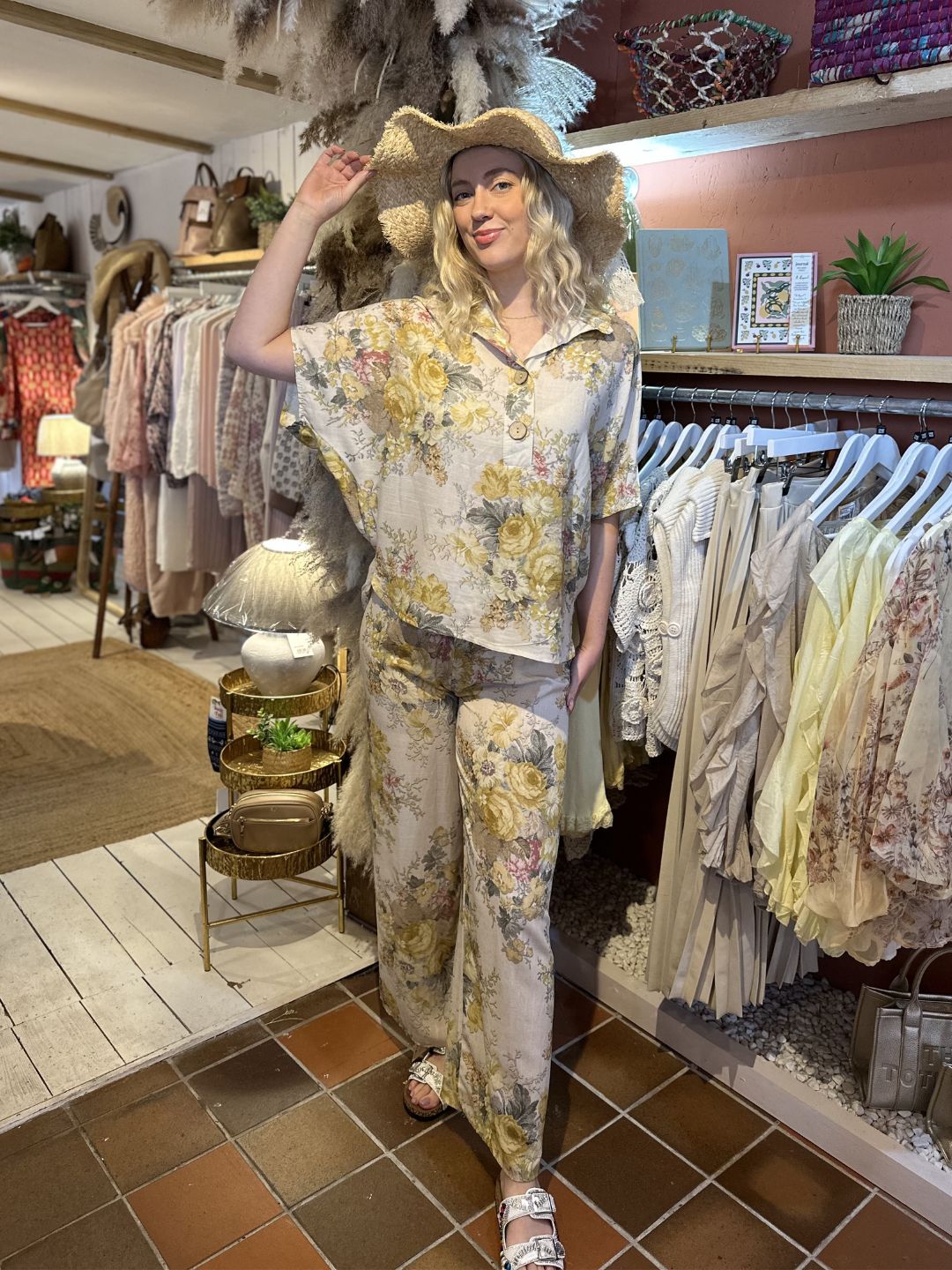 A woman in a floral co-ord set with wide-leg trousers, a wide-brimmed hat, and sandals poses in a boutique filled with hanging clothes and assorted decorative items.