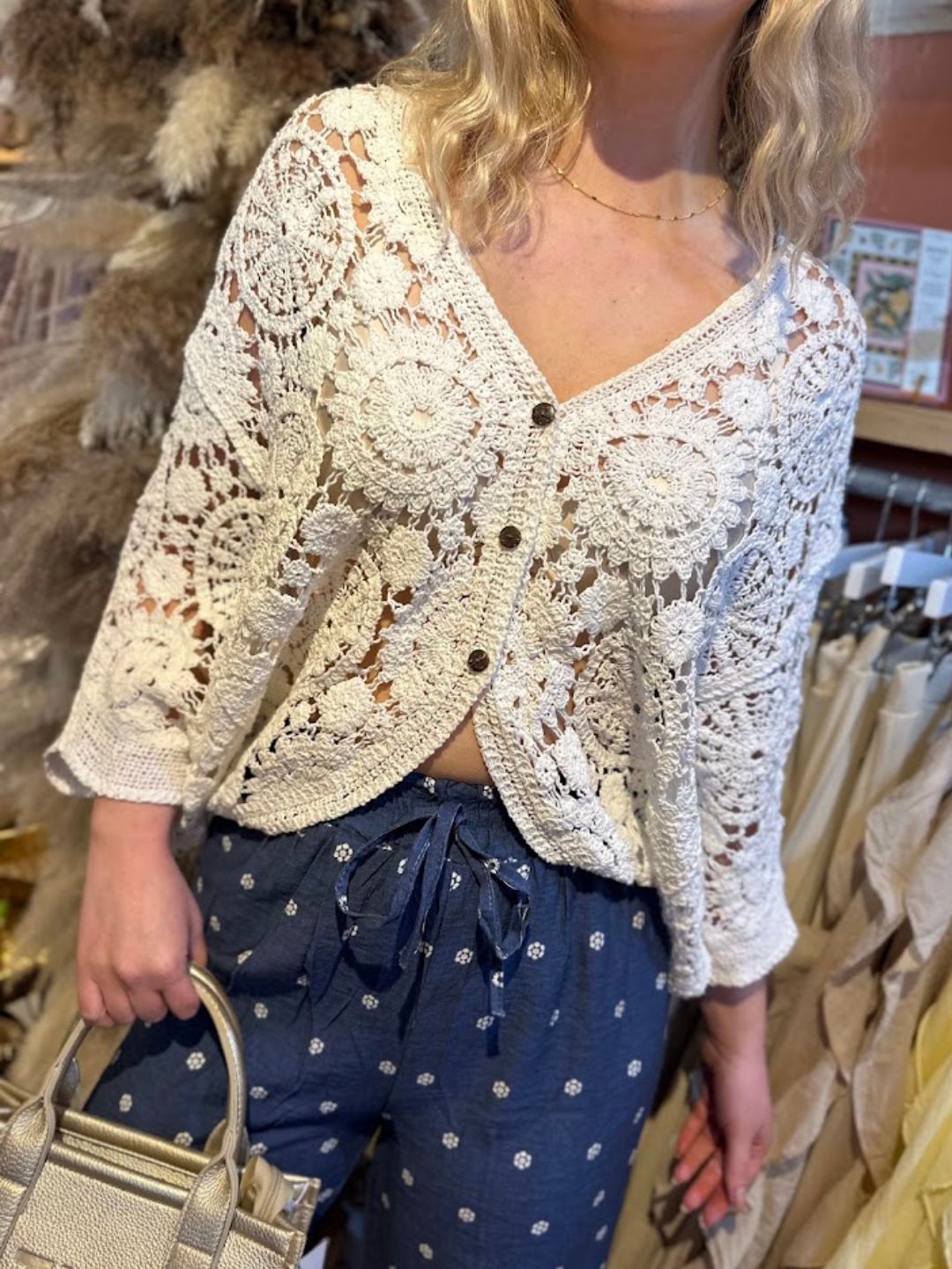A person wearing a white open-front crochet cardigan with floral patterns, blue drawstring trousers featuring small white designs, and carrying a beige handbag.