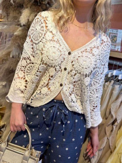 A person wearing a white open-front crochet cardigan with floral patterns, blue drawstring trousers featuring small white designs, and carrying a beige handbag.