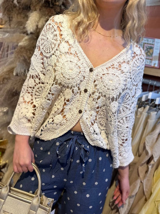 A person wearing a white open-front crochet cardigan with floral patterns, blue drawstring trousers featuring small white designs, and carrying a beige handbag.