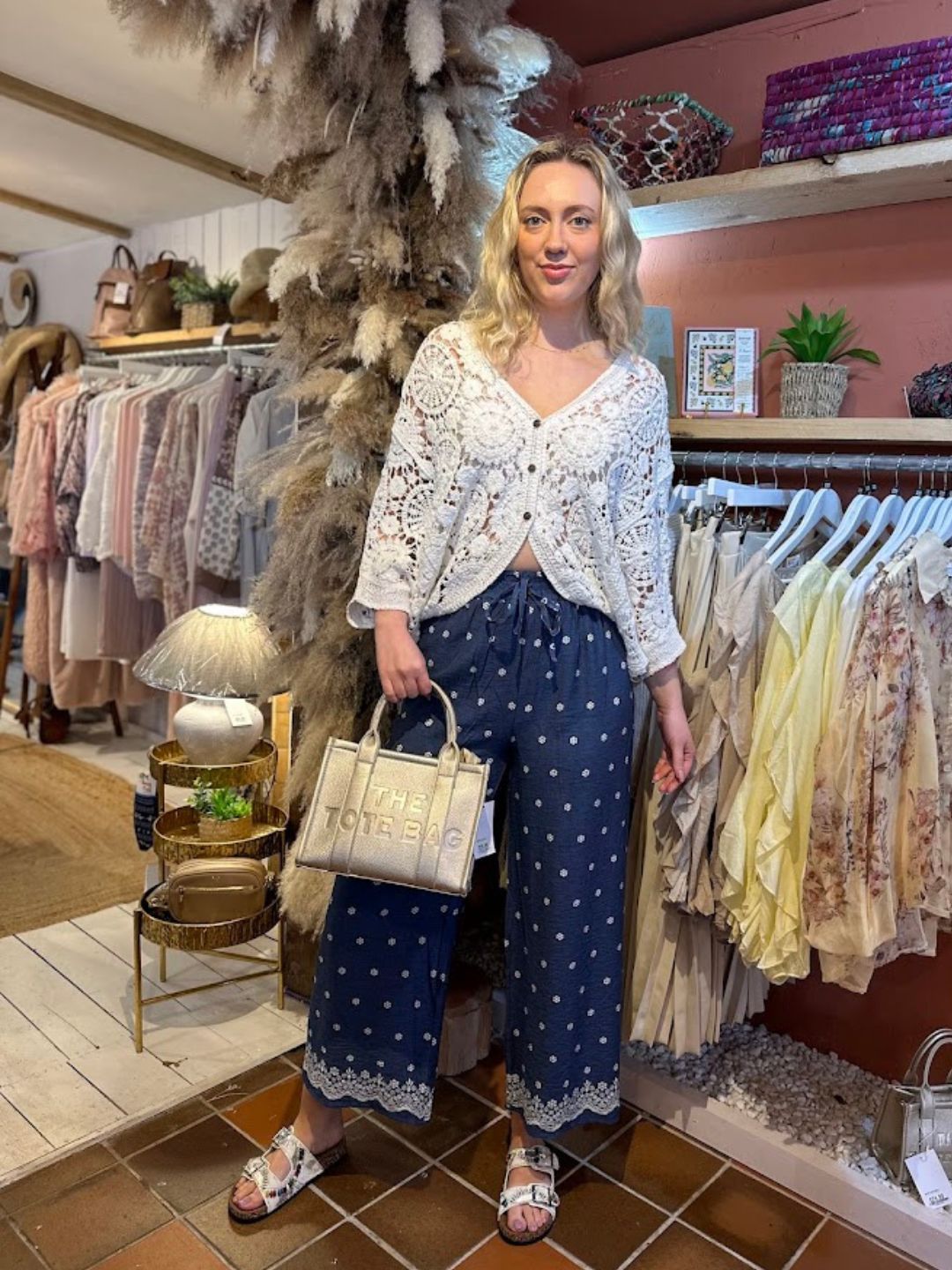A woman stands in a boutique wearing a white crochet cardigan over a lace top, navy patterned wide-leg trousers, sandals, and holding a beige tote bag. She is surrounded by clothing displays and décor.