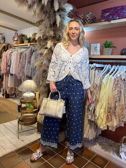 A woman stands in a boutique wearing a white crochet cardigan over a lace top, navy patterned wide-leg trousers, sandals, and holding a beige tote bag. She is surrounded by clothing displays and décor.