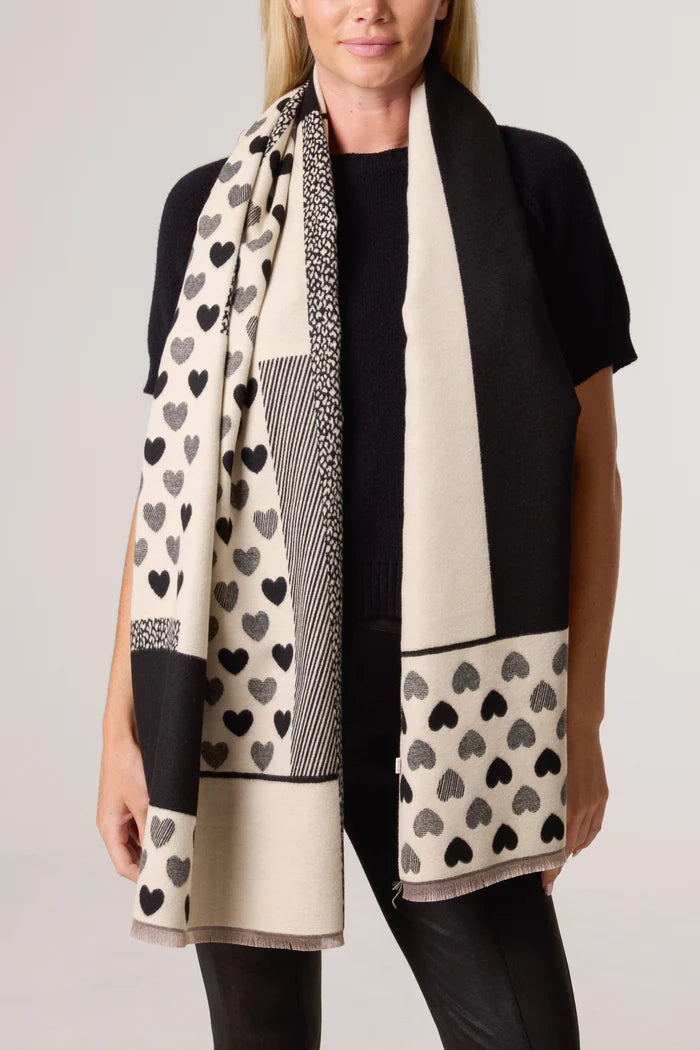 A woman in a black top and trousers models a reversible vegan heart-print scarf, showcasing striking black, cream, and geometric patterns for a bold statement.