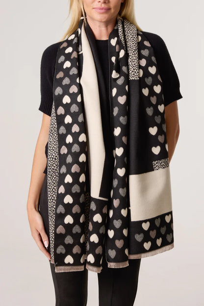 A woman wears a black short-sleeved top and a long reversible vegan heart print scarf in black and cream, featuring heart motifs and small geometric sections.