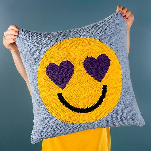 Person holding a Smile Knitted Cushion, a square polyester cushion with a yellow smiley face featuring heart-shaped eyes and a black smile—ideal for adding fun to your home décor against a light blue background.