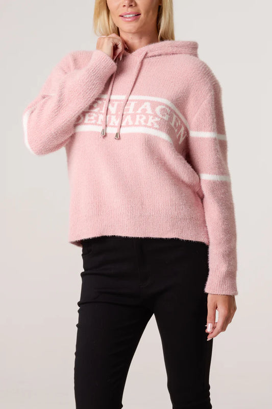 A woman wearing a baby pink drawstring hoodie with white stripes and lettering, paired with black trousers, stands against a plain background.