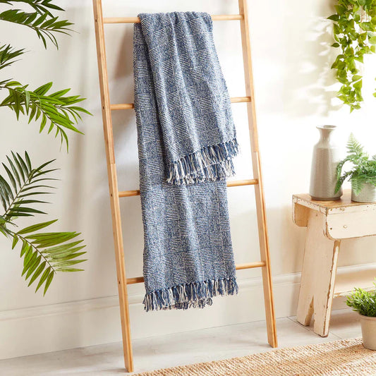 A blue and white fringed woven throw, crafted from recycled bottles, draped over a wooden ladder and surrounded by green plants and simple décor in a bright room.