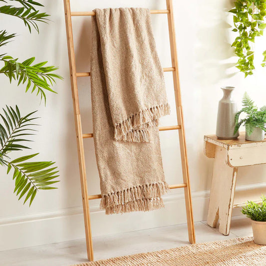 A beige Woven Cross Hatch Throw, crafted from recycled bottles, hangs on a wooden ladder beside houseplants and a small bench in a bright, cosy room.