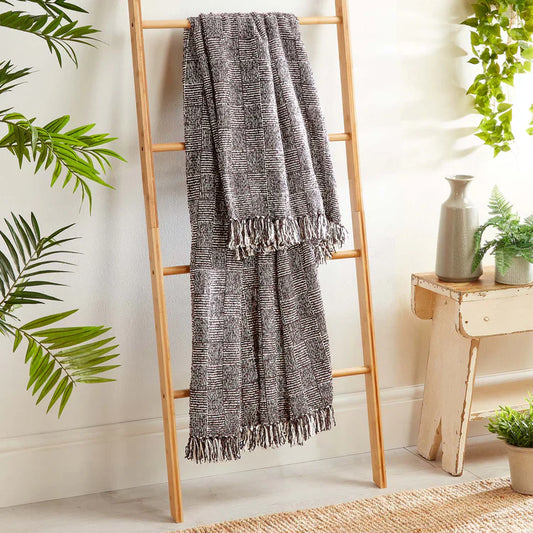 A grey and white woven crosshatch throw with fringe hangs on a wooden ladder beside a bench and potted plants in a bright room.