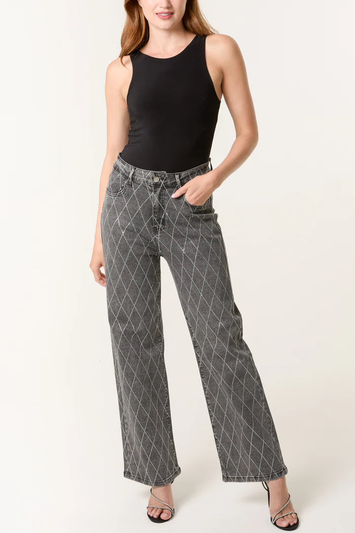 A woman in a sleeveless black top, stretch denim grey wide-leg jeans with a white criss-cross pattern, and black heeled sandals stands with one hand in her pocket.