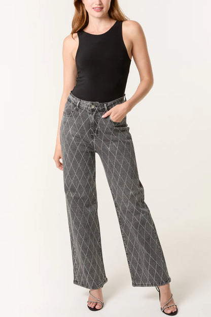 A woman in a sleeveless black top, stretch denim grey wide-leg jeans with a white criss-cross pattern, and black heeled sandals stands with one hand in her pocket.