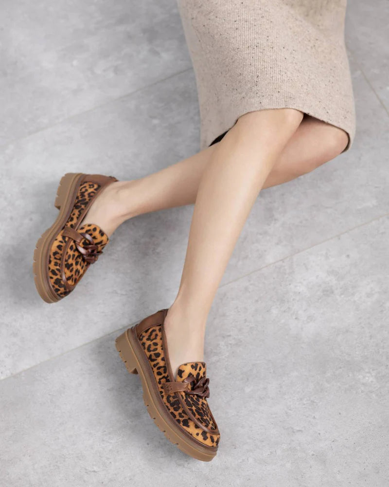 A person wearing a beige knitted skirt and leopard print women’s moccasins with thick brown soles sits on a light grey stone floor.