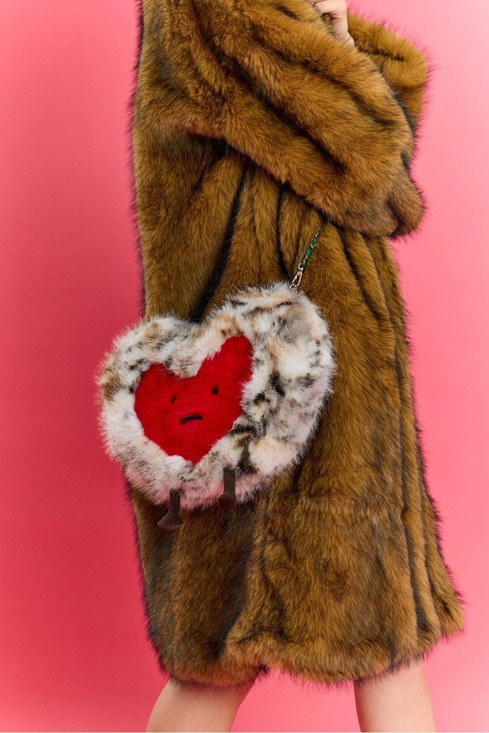 Person in a brown faux fur coat holds a faux fur heart-shaped bag featuring a red face design, set against a pink background.