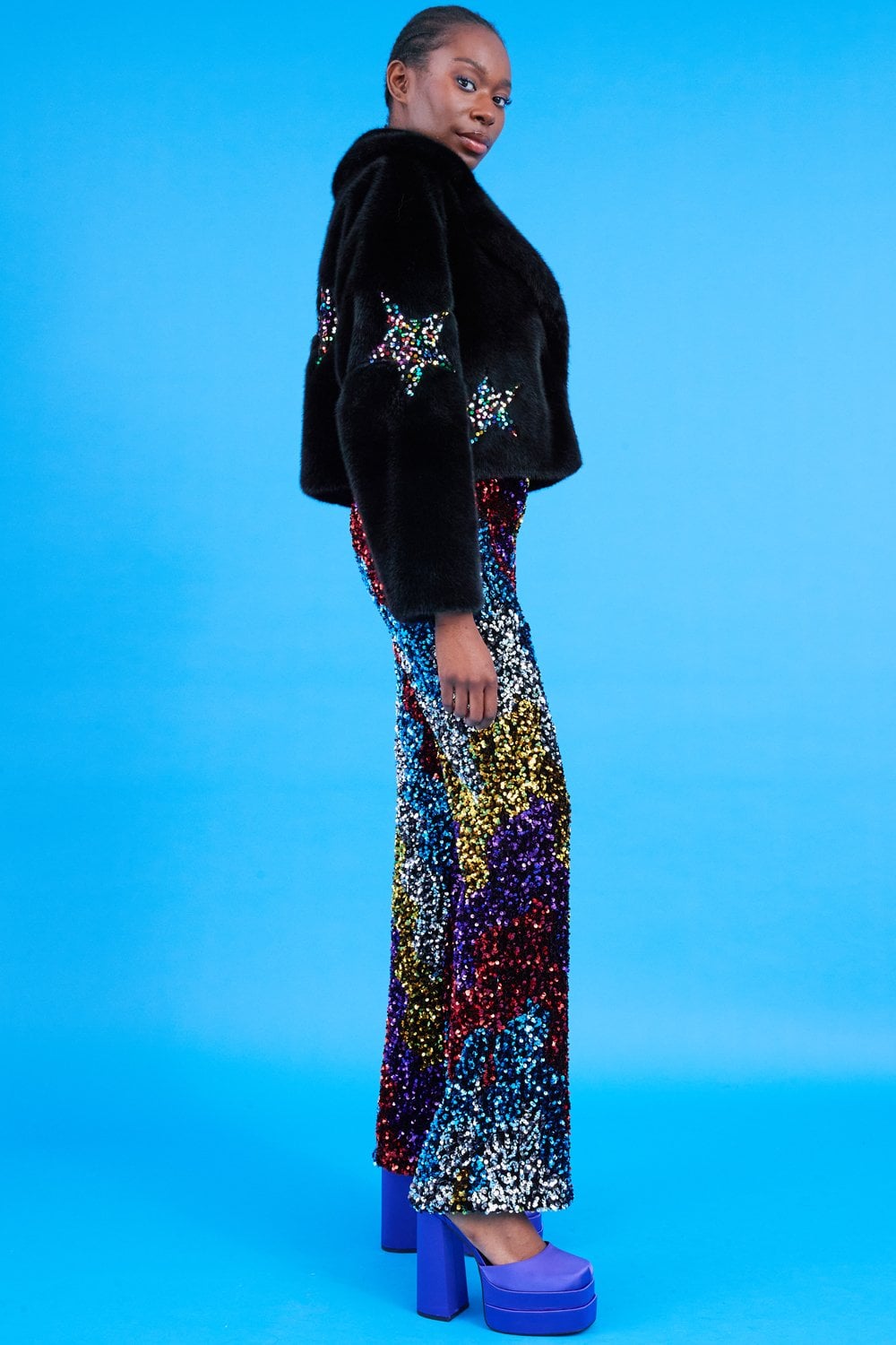 Person standing against a blue background, dressed in a black bamboo jacket adorned with sequin star details, vibrant sequin trousers, and purple platform shoes.