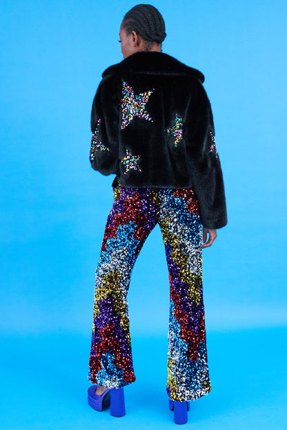 Person standing against a blue background, wearing a Black Bamboo Jacket with faux fur and sequin star details, paired with high-waisted, multicoloured sequin trousers and striking blue platform shoes.