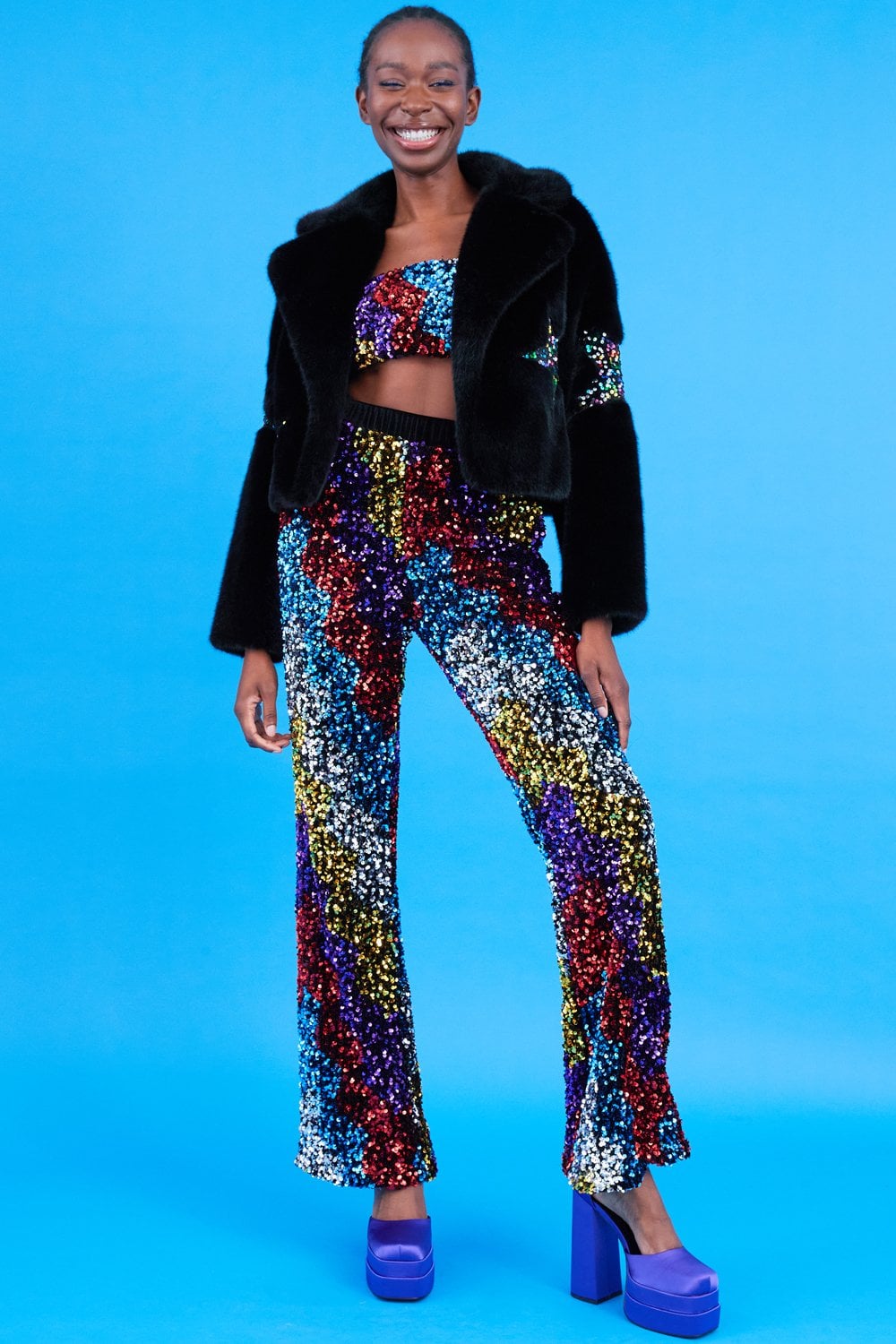 A person smiles in a black faux fur jacket, sequin star crop top and trousers set featuring multicoloured patterns, and purple platform heels against a bright blue background.