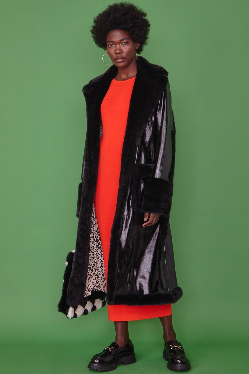A person stands against a green background, dressed in a long black faux leather trench coat with faux fur detailing over a bright red dress and black shoes.