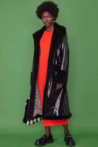 A person stands against a green background, dressed in a long black faux leather trench coat with faux fur detailing over a bright red dress and black shoes.