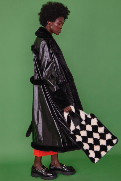 A person wearing a black faux leather trench coat with faux fur detailing and a leopard print lining holds a black and white chequered bag, standing against a green background.