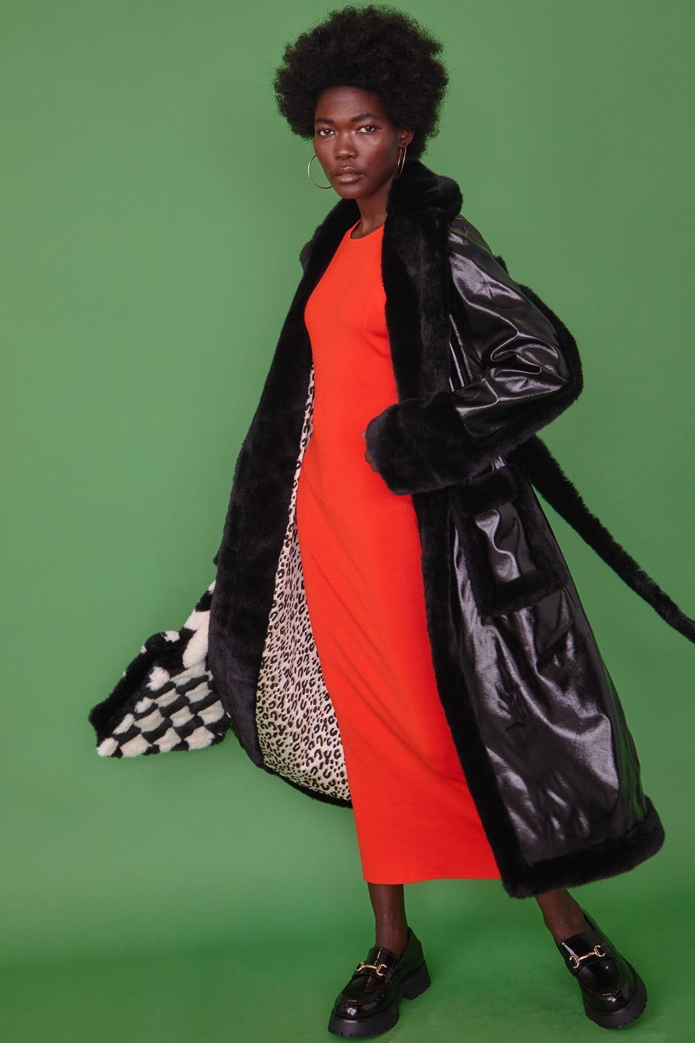 A woman in a long red dress and black faux leather trench coat stands against a green background, showing the coat’s chequered and leopard print lining.