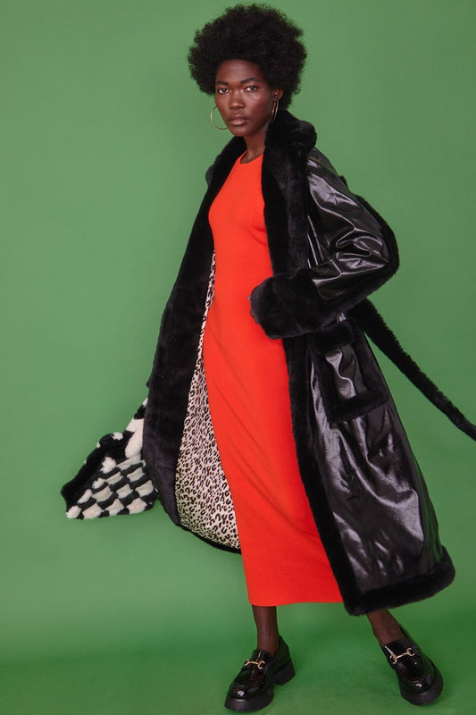 A woman in a long red dress and black faux leather trench coat stands against a green background, showing the coat’s chequered and leopard print lining.