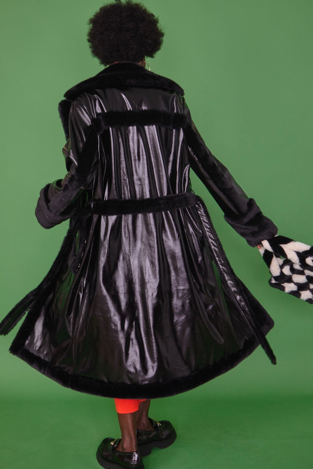 Person wearing a black faux leather trench coat with faux fur detailing and red trousers stands against a green background, facing away from the camera, holding a black and white patterned bag.