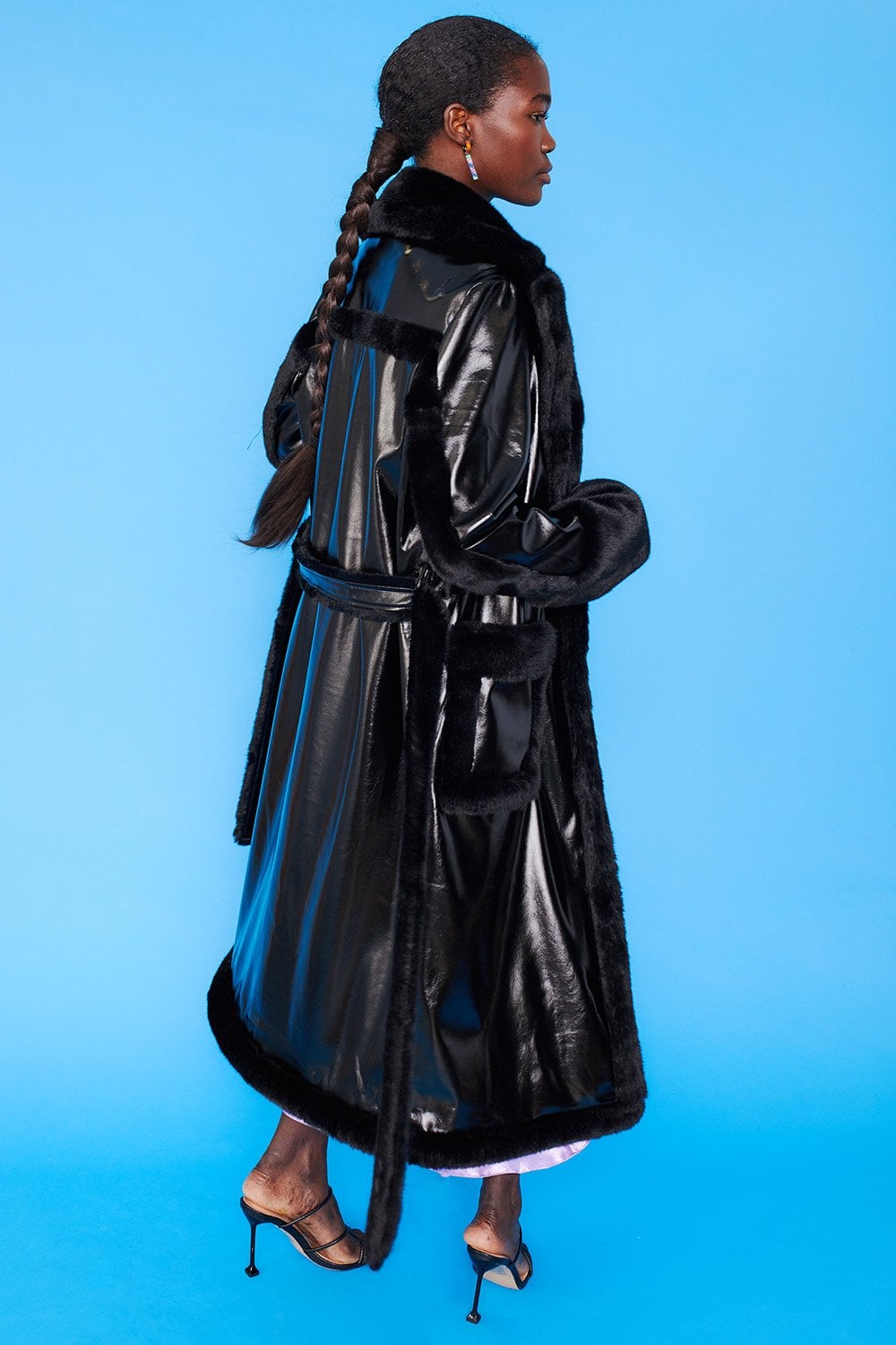 A person in a black faux leather trench coat with leopard print lining and heeled sandals stands against a solid blue background.